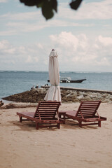 Two empty wooden lounge chairs and a closed parasol on a sandy tropical beach overlooking the calm ocean