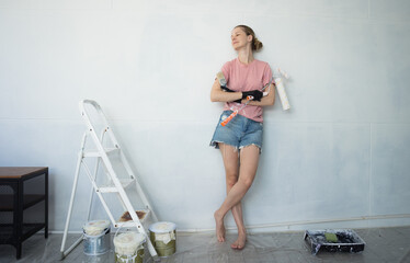 Young woman painting indoors wall with roller using stepladder