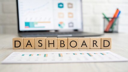 Wooden cubes spelling "DASHBOARD" with blurred analytics screen behind
