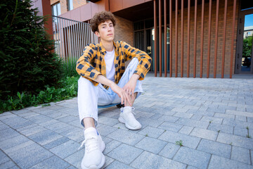 A young man in a yellow checkered shirt sits on a blue skateboard with his legs crossed against a...