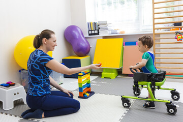 Caring therapist assists cute young boy patient as he practices walking with rehab walker in...