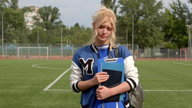Portrait of upset beautiful blonde high school female student with textbook and digital tablet wearing uniform and varsity jacket, expressing disappointment and sadness after failed exam outdoors.