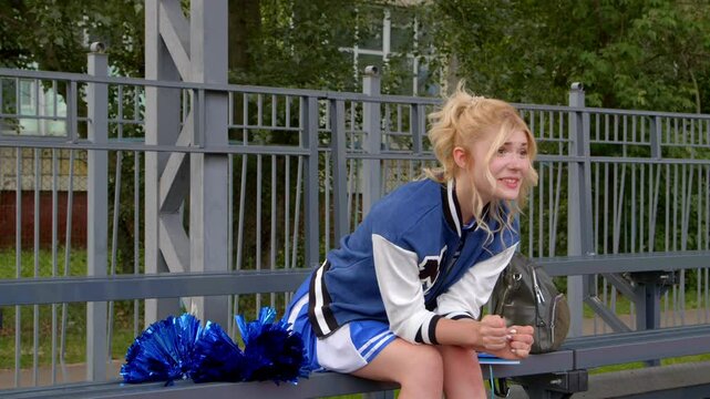 Fully in engrossed in game charming blonde high school girl wearing uniform and letterman jacket, emotionally celebrating successful play of sport team and clapping hands while sitting on bleacher.