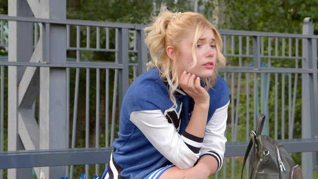 Portrait of upset attractive blonde high school female student in varsity jacket and uniform sitting on bleacher , being disappointed and depressed after failed exam.