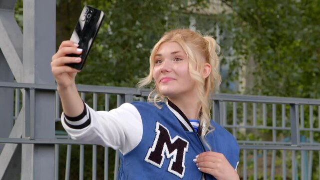 Portrait of beautiful blonde popular teenage girl in varsity jacket, taking selfie shot on mobile phone, making funny faces and gesture , expressing carefree mood and happiness outdoors.