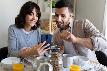 Happy couple eating healthy breakfast at home looking at mobile phone