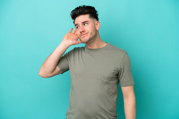 Young caucasian man isolated on blue background listening to something by putting hand on the ear