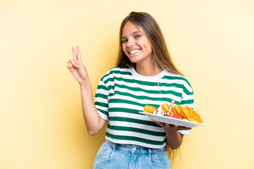 Young caucasian woman holding waffles isolated on yellow background smiling and showing victory sign
