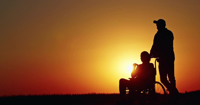 Silhouette of a man gently pushing a woman in a wheelchair during sunset, symbolizing care, support, and companionship in a peaceful rural landscape.