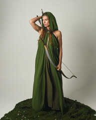 Full length portrait of female model figure wearing green fantasy fae hooded cloak crown headdress costume, holding archery bow weapon in hunting pose, standing on mossy floral studio background. 