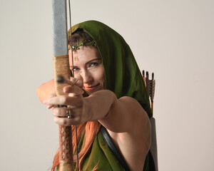 close up portrait of female model figure wearing green fantasy fae hooded cloak crown headdress costume, holding archery bow  weapon in hunting pose, shooting camera. isolated studio background