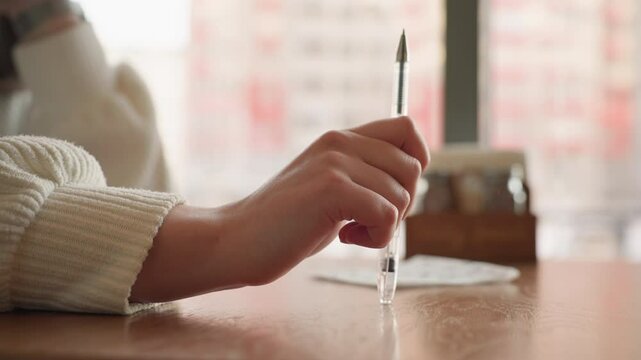 Close up hand holding pen and gently tapping it on wooden table beside napkin in soft natural indoor light suggesting thoughtful mood, waiting moment