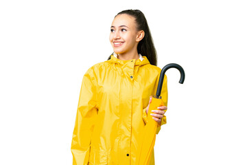 Young Arabian woman with rainproof coat and umbrella over isolated chroma key background looking to the side and smiling