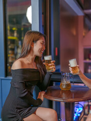 Beautiful young woman enjoying beer with friend at bar, smiling and toasting together. Nightlife, celebration, friendship and happiness concept.