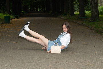 A young woman in a denim outfit lies on her back on a park path, legs raised playfully. She smiles...