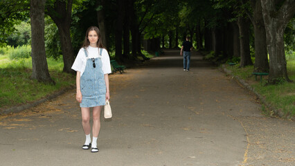 A young woman stands confidently on a wide path surrounded by tall trees in a city park. A person...