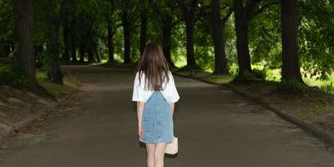 A young woman strolls down a serene path surrounded by lush trees in a peaceful park. The sunlight filters through the leaves, creating a calm atmosphere