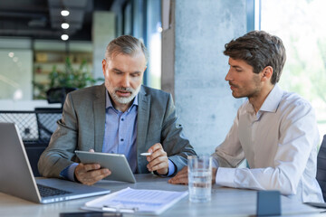 Mature businessman using a digital tablet to discuss information with a younger colleague in a...