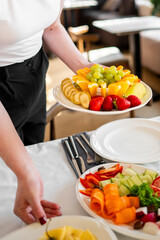 Close-up of a person serving a plate with fresh fruits including bananas, grapes, oranges, and strawberries, alongside a plate with assorted vegetables on a table indoors.