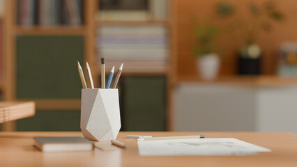 A 3D-rendered geometric white pencil holder with angular faceted design sits on a wooden desk, containing colored pencils and pens, surrounded by notebooks and office supplies in natural light.