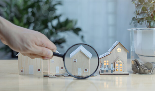Hand holding magnifying glass inspecting house model. Concept of real estate selection, property evaluation, home buying decisions, and investment in residential housing.