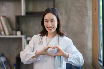 A female doctor is looking at the camera, making a heart gesture with both hands.