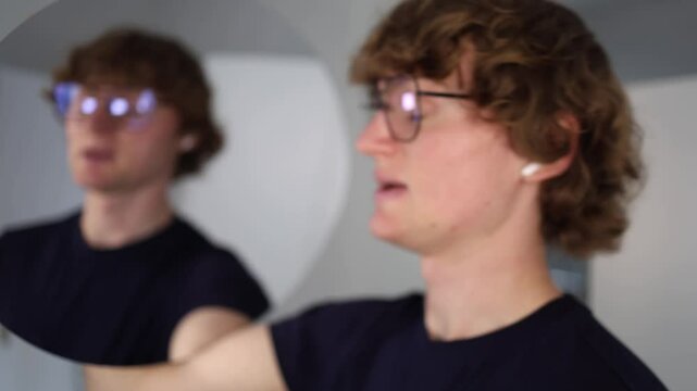 Man with glasses puffs his cheeks and stares into the mirror with a tense expression as if gathering thoughts or preparing for something serious alone in a quiet modern interior