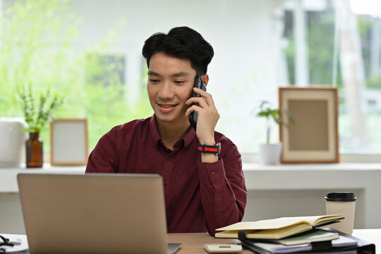 A smiling young Asian man multitasking between a laptop and a phone call in a bright, modern office space - Powered by Adobe