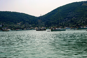 Sailboats anchored in a calm bay surrounded by lush green hills and coastal village, Mediterranean...