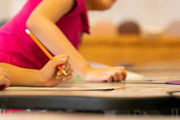 Young elementary school student in pink shirt grips a number 2 pencil while focused on writing assignment at wooden desk in modern classroom setting. 