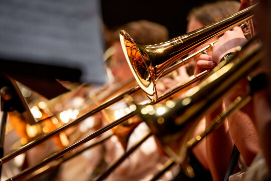 Close-up view of high school band students performing with brass instruments, including trumpets and trombones, during a school concert. The image captures the golden metallic shine of the instruments