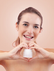 Smiling young woman with makeup holding a plastic cosmetic jar of face cream while make a heart...