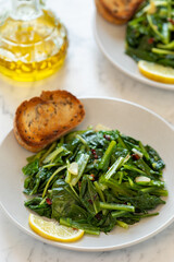 Roman-style sautéed chicory greens (Cicoria Ripassata alla Romana) served on a plate with toasted bread and a slice of lemon. A bottle of olive oil and a second plate are in the background.