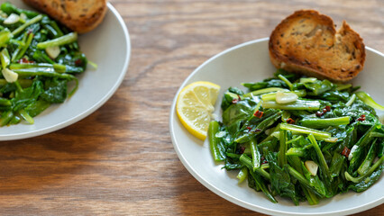 Two plates with Roman-style chicory greens sautéed in olive oil (Cicoria Ripassata alla Romana) served with toasted bread and a slice of lemon on a brown wooden table.