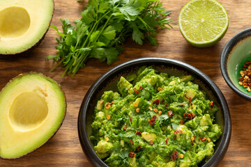 Homemade guacamole in a bowl on a brown wooden table with the main ingredients shown: avocado cut in half, lime, and a bunch of coriander.