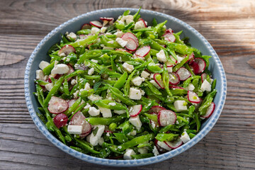 Platter with sugar snap pea and radish salad with torn mint leaves and aged ricotta cheese on a rustic wooden table.