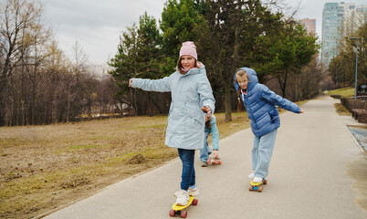 Fototapeta premium Teen girls ride skateboards on city path in winter autumn park. Jackets and jeans reflect urban youth style. Street sport improves balance, movement, coordination, friendship and outdoor fun for kids