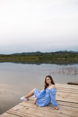 Girl in blue dress sitting on pier on wooden planks near lake