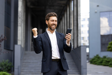 A businessman in a suit celebrates success while looking at his phone. He is standing outdoors near a building.
