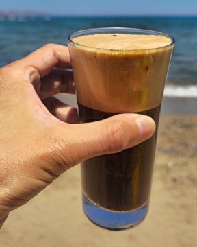Iced freddo espresso with thick crema held in hand at Greek beach, summer sunlight and deep blue sea in background create the perfect vacation coffee moment. Kissamos, Greece, July 13, 2025