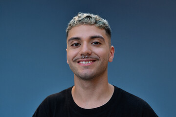 Smiling young man with bleached curly hair and septum piercing posing confidently against a plain...