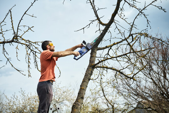 Young man cutting trees using electrical chainsaw in motion at his backyard
