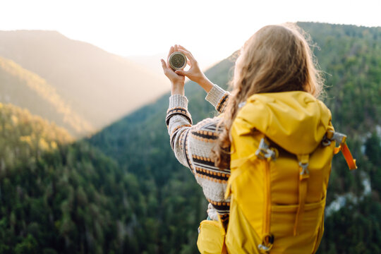 Close up of young female traveler hands with yellow backpack holding compass against mountain landscape. Woman with compass looking direction standing on cliff. Navigation, travel and hiking concept. - Powered by Adobe