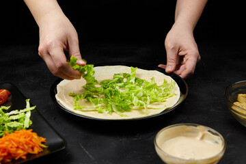 Hands of a person adding fresh green lettuce to a tortilla on a black plate, surrounded by colorful vegetables and a bowl of sauce, showcasing healthy meal preparation