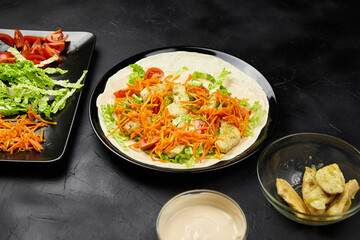 Freshly prepared tortilla wrap filled with grilled chicken, lettuce, carrots, and tomatoes, served on a black surface with additional ingredients in the background