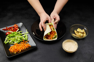 Hands of a person preparing a fresh vegetable wrap with colorful ingredients on a black surface, showcasing culinary skills and healthy eating concepts
