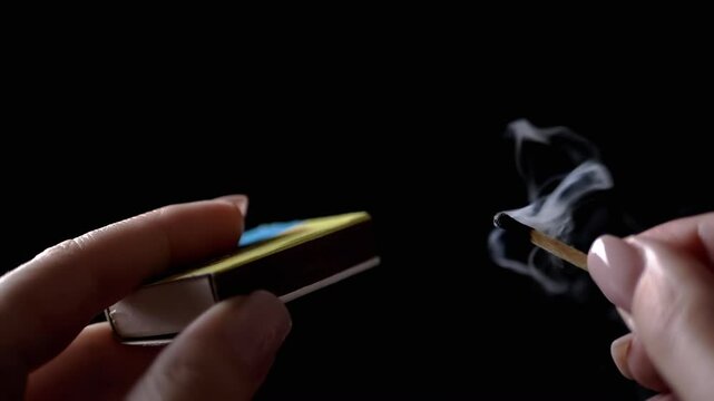 Blowing Out Burning Match in Dark Room. Female Hands. Woman Holding Burning Match with Matchbox, Then Blowing It Out. Puffs of smoke, fire. Empty space. Black background. Slow motion.