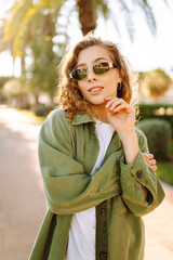 Cheerful woman with curly hair and sunglasses enjoys a sunny day on an alley with palm trees. A...