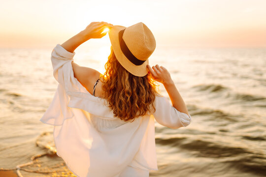 Happy woman in a hat having fun on a sandy beach in the rays of sunset. Young woman enjoying the seascape and relaxing outdoors. Concept of nature, weekend and freedom.