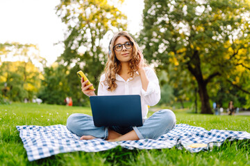 Cheerful woman sits on a blanket in a sunlit park with a laptop and listens to music on wireless...